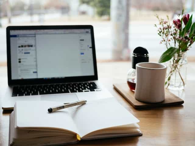 An open notebook lying flat on a table in front of a laptop next to a coffee mug and flower vase.